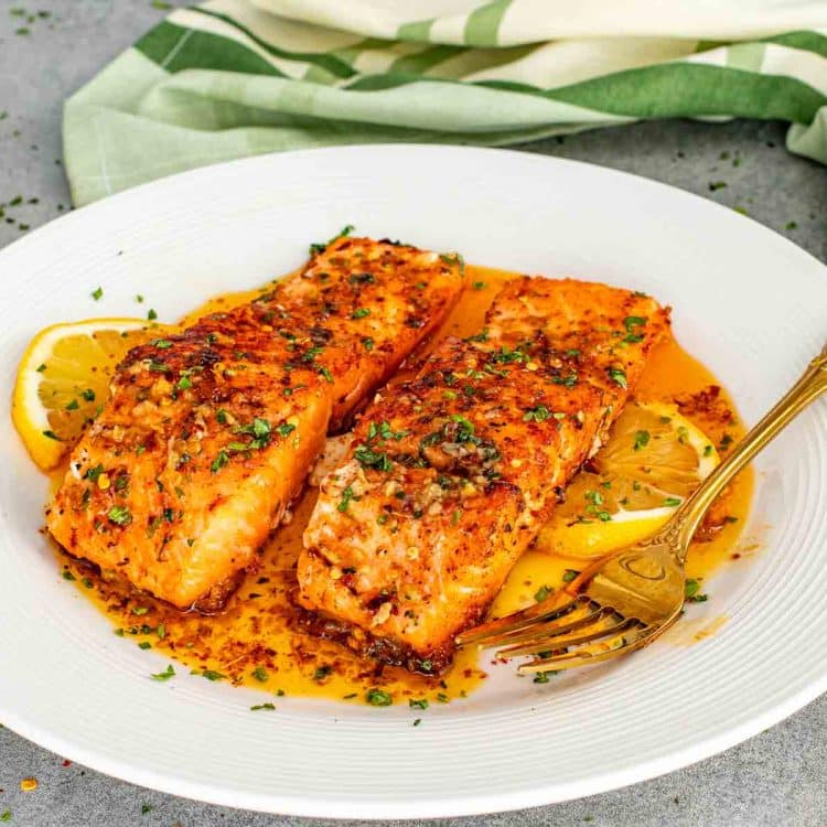 Two salmon fillets served with lemon slices and spooned cowboy butter sauce on white dinner plate.