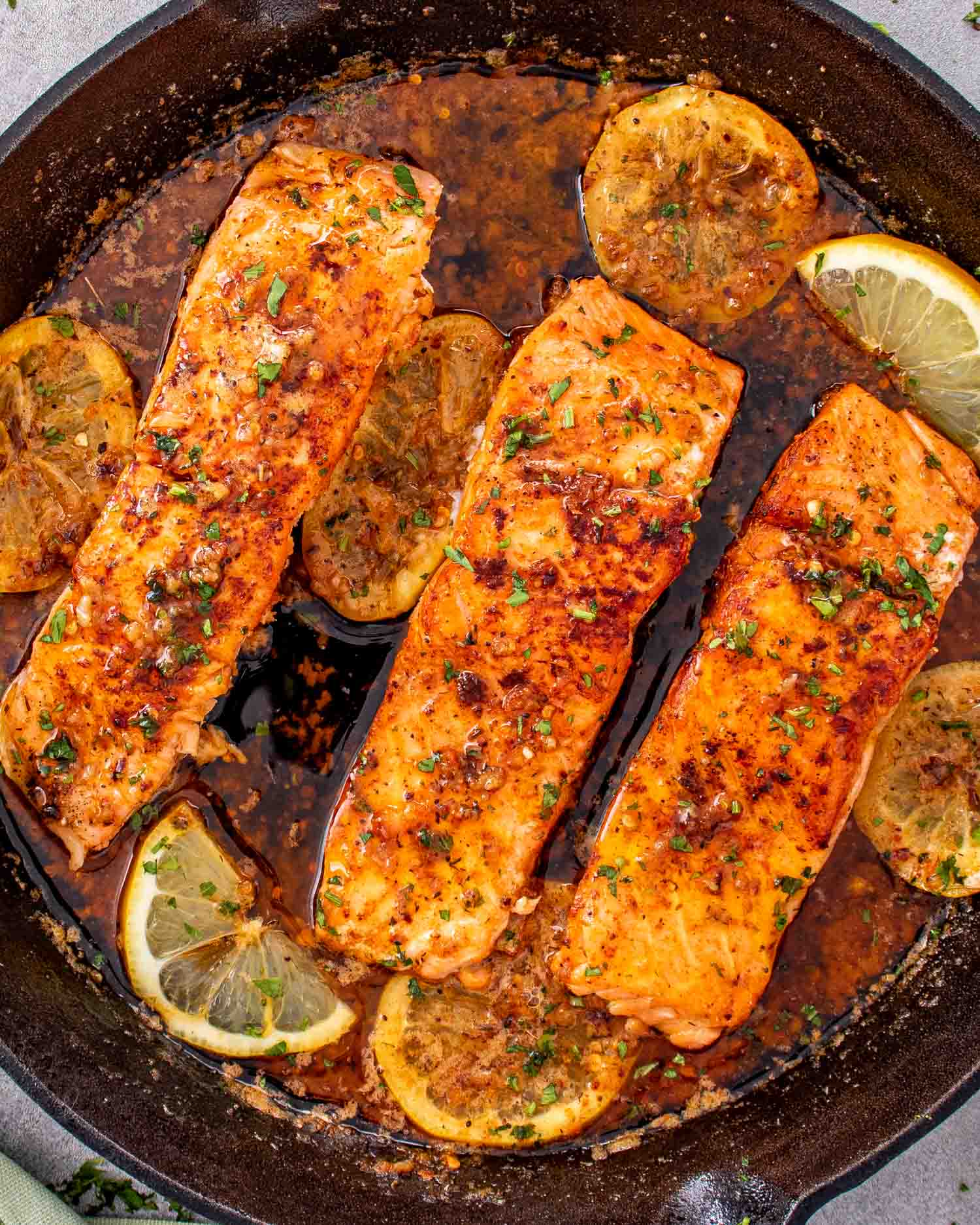 Pan seared salmon fillets simmering in cowboy butter sauce with lemon slices in cast iron skillet.