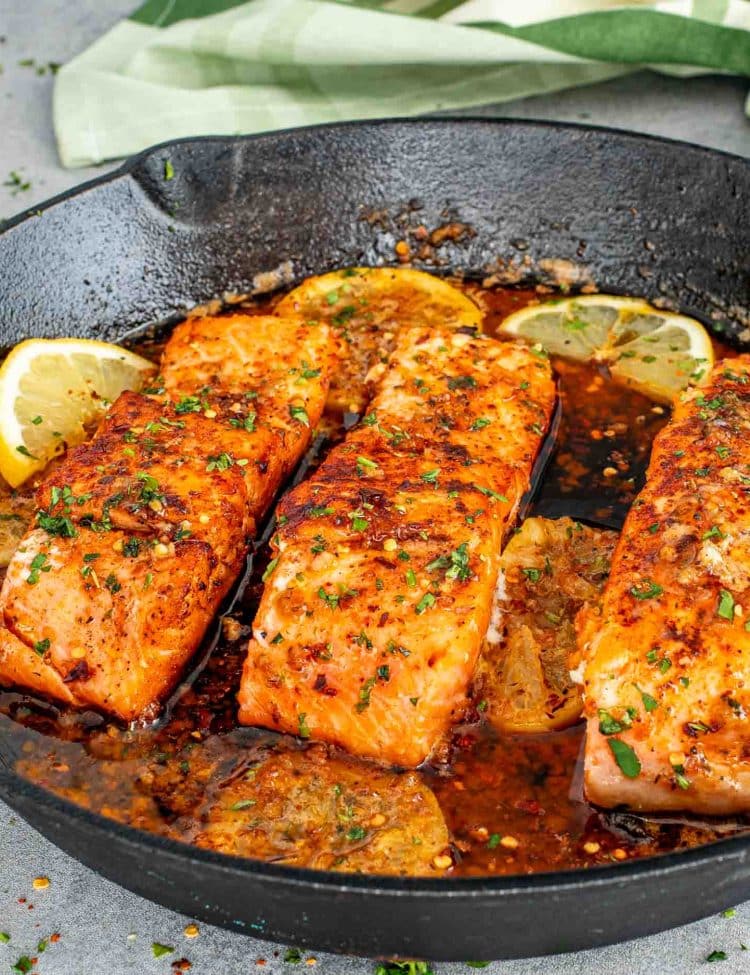 Skillet cowboy butter salmon fillets searing in garlic butter sauce with lemon slices and herbs in cast iron pan.