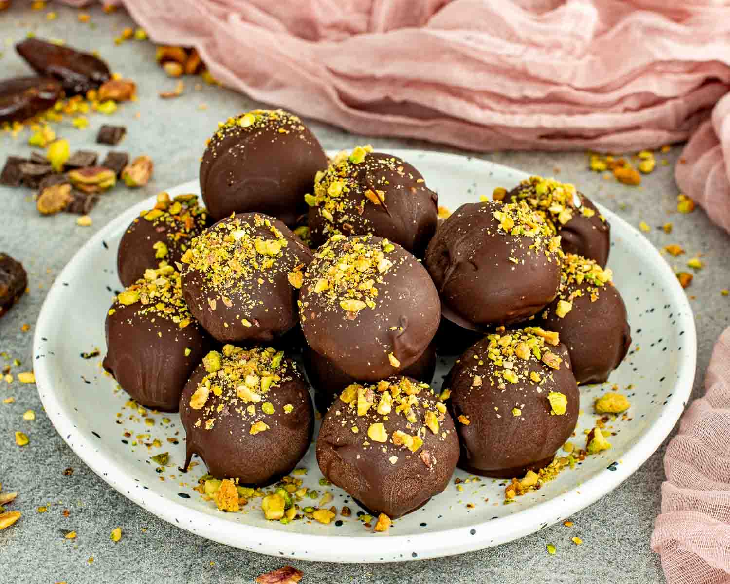 Homemade pistachio date chocolate balls topped with crushed pistachios on a white plate.