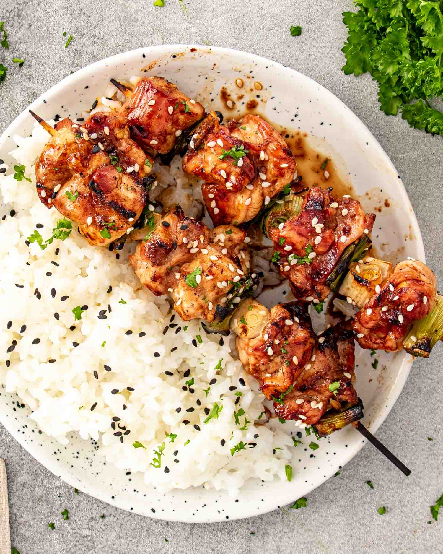 A top down view of two grilled chicken skewers served over a bed of white rice in a white bowl and garnished with black sesame seeds and fresh herbs.