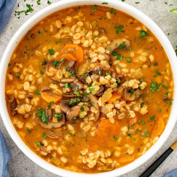 Overhead view of mushroom barley soup with barley, mushrooms, carrots, and herbs in a rich broth, served in a white bowl.