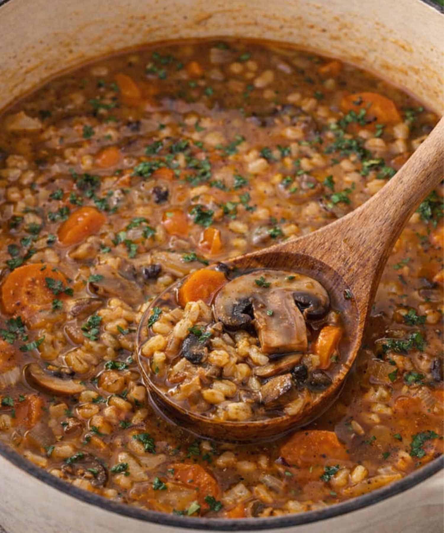 Mushroom barley soup cooking in a pot with wooden spoon, showing tender barley, mushrooms, carrots, and thick savory broth.