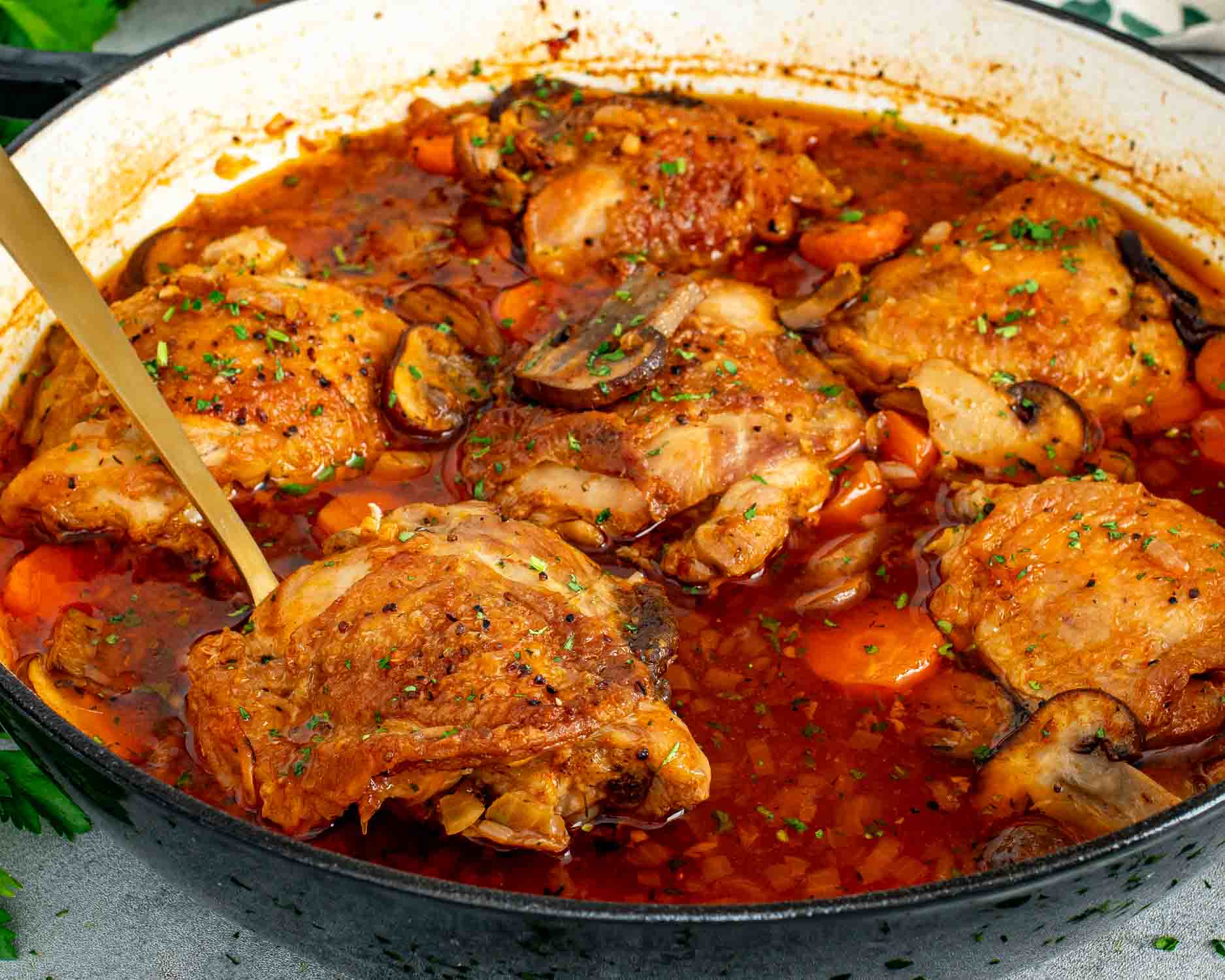 Close up angled view of 6 braised chicken thighs in a deep skillet with sliced mushrooms, carrots, and rich red braising liquid garnished with fresh parsley and a gold serving spoon.