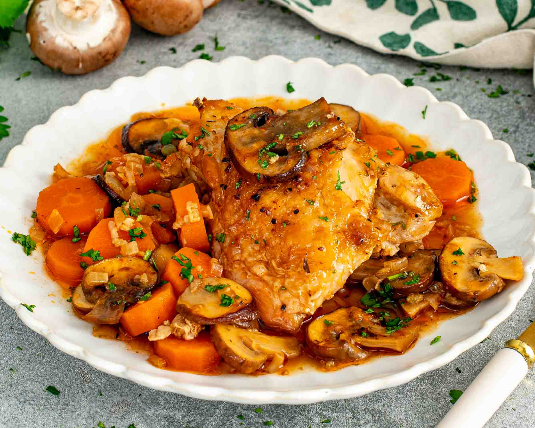A golden braised chicken thigh in a white scalloped bowl surrounded by sliced mushrooms, chunky carrots, onion, and savory braising liquid garnished with fresh parsley and a gold fork.