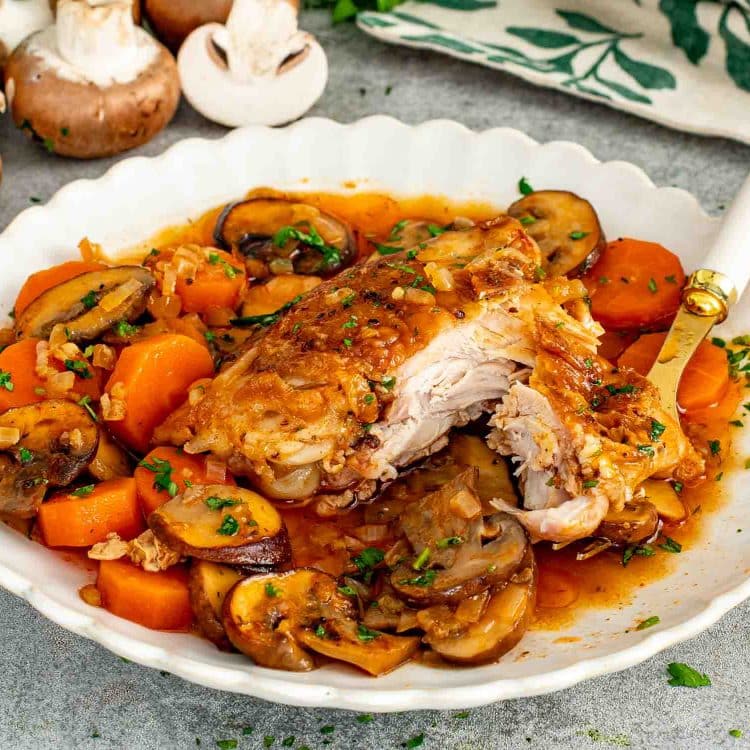 A braised chicken thigh pulled apart to show tender meat in a white scalloped bowl with sliced mushrooms, carrots, onion, and rich braising sauce garnished with fresh parsley.
