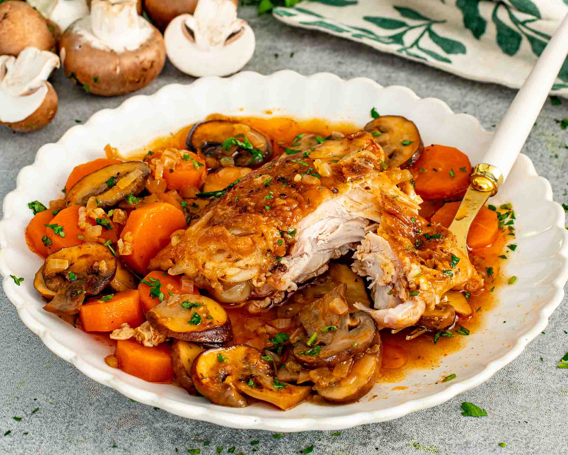 A braised chicken thigh pulled apart to show tender meat in a white scalloped bowl with sliced mushrooms, carrots, onion, and rich braising sauce garnished with fresh parsley.