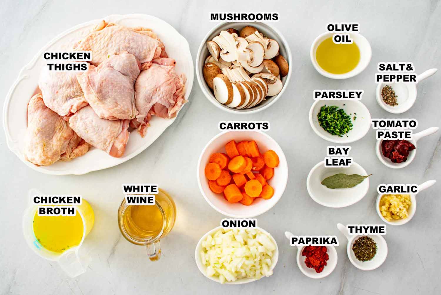 Overhead flat lay of braised chicken ingredients including bone in skin on chicken thighs, sliced mushrooms, carrots, onion, white wine, chicken broth, tomato paste, garlic, bay leaf, paprika, thyme, parsley, olive oil, salt, and pepper.