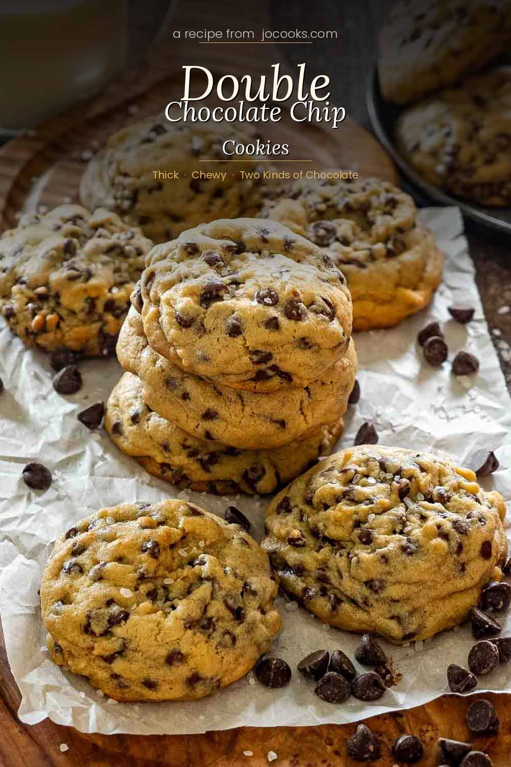 Thick double chocolate chip cookies piled on a wood board with parchment paper, scattered dark and semisweet chocolate chips and flaky sea salt, with script text overlay at the top.