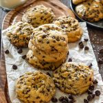 Stack of three double chocolate chip cookies on parchment paper on a wood serving board, surrounded by more cookies, scattered chocolate chips, flaky sea salt, and a glass of milk in the background.