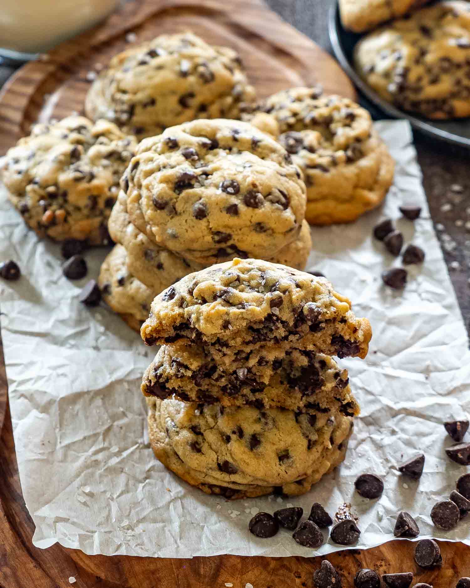 A double chocolate chip cookie broken in half and stacked to show the soft gooey interior packed with melted dark and semisweet chocolate chips, with more cookies and sea salt on parchment paper in the background.