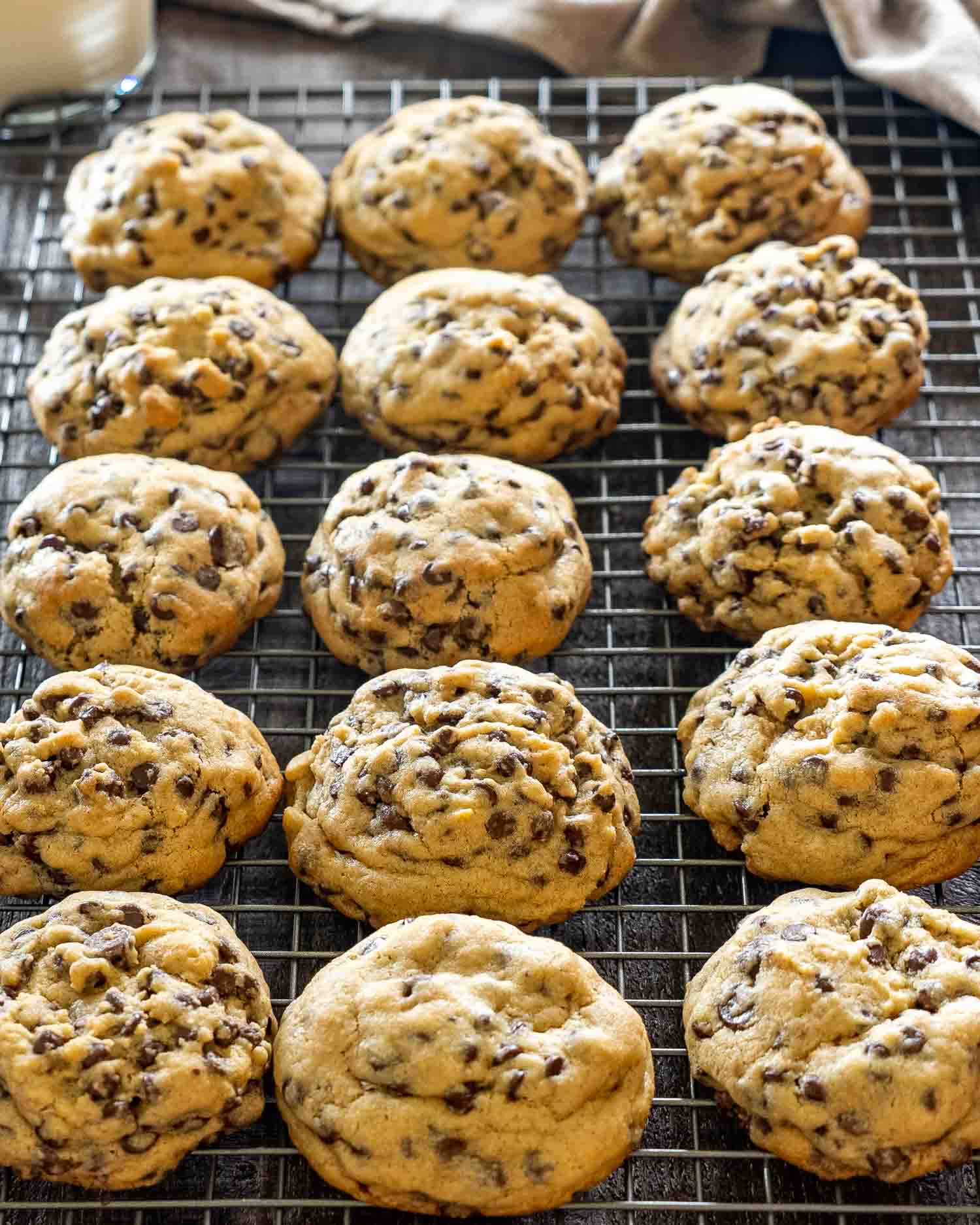 Overhead view of fifteen thick double chocolate chip cookies cooling on a wire rack on a dark wood surface, showing the golden edges and chocolate chip studded tops.