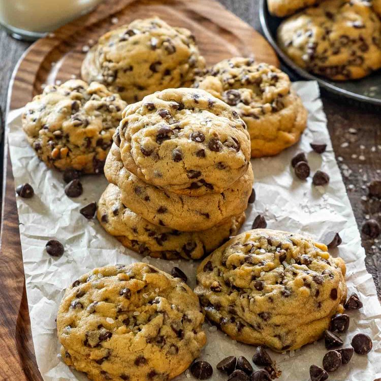 Stack of three double chocolate chip cookies on parchment paper on a wood serving board, surrounded by more cookies, scattered chocolate chips, flaky sea salt, and a glass of milk in the background.