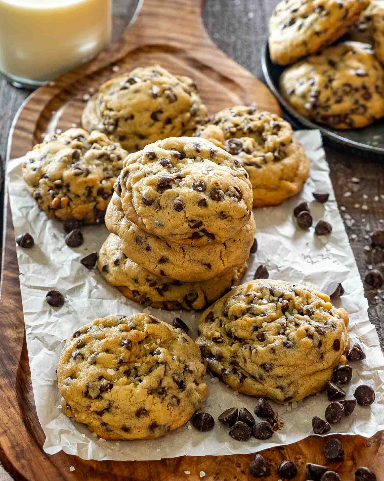 Stack of three double chocolate chip cookies on parchment paper on a wood serving board, surrounded by more cookies, scattered chocolate chips, flaky sea salt, and a glass of milk in the background.