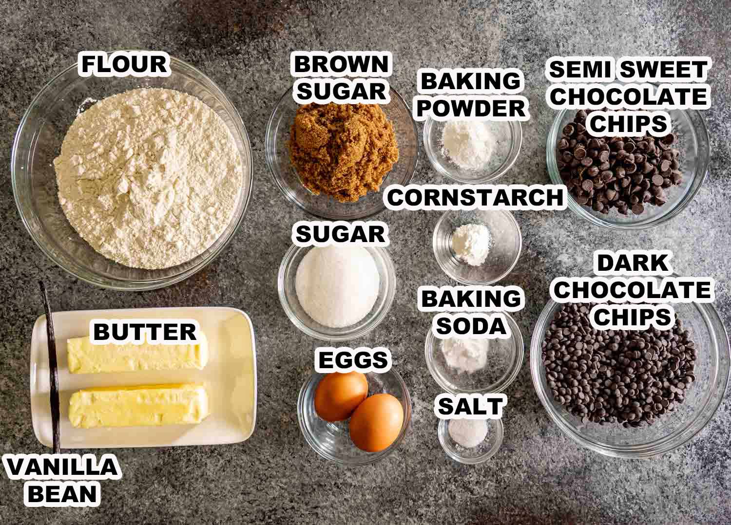 Overhead flat lay of double chocolate chip cookie ingredients including flour, dark brown sugar, butter, vanilla bean, eggs, cornstarch, baking powder, baking soda, salt, semisweet chocolate chips, and dark chocolate chips, each labeled on a gray surface.