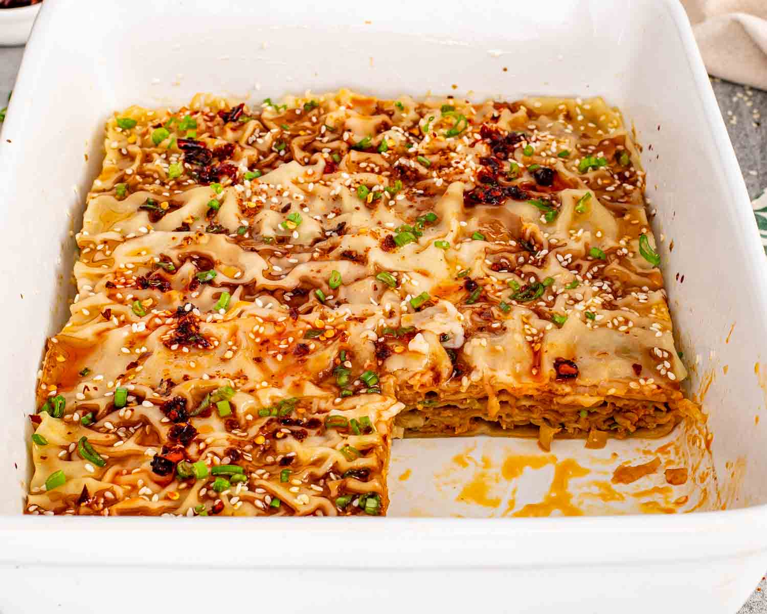 Top view of a white casserole dish filled with golden brown dumpling lasagna, garnished with green onions and sesame seeds.