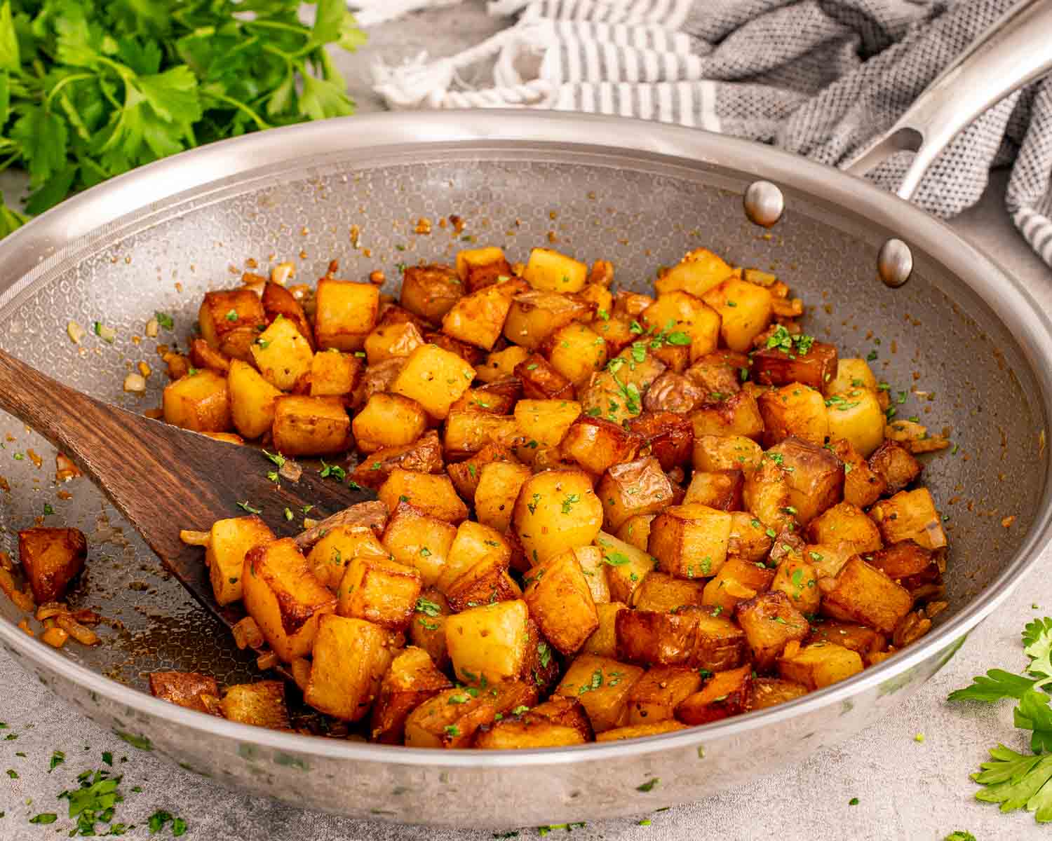 Golden pan fried home fries with caramelized onions and parsley in large skillet with wooden spatula on neutral background.