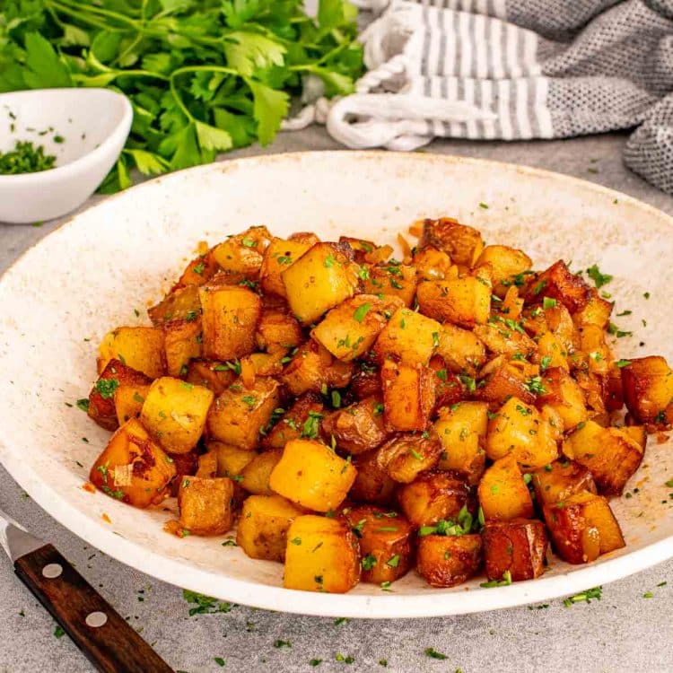 Close up rustic bowl filled with crispy home fries, caramelized onions, parsley, and golden browned potato cubes.