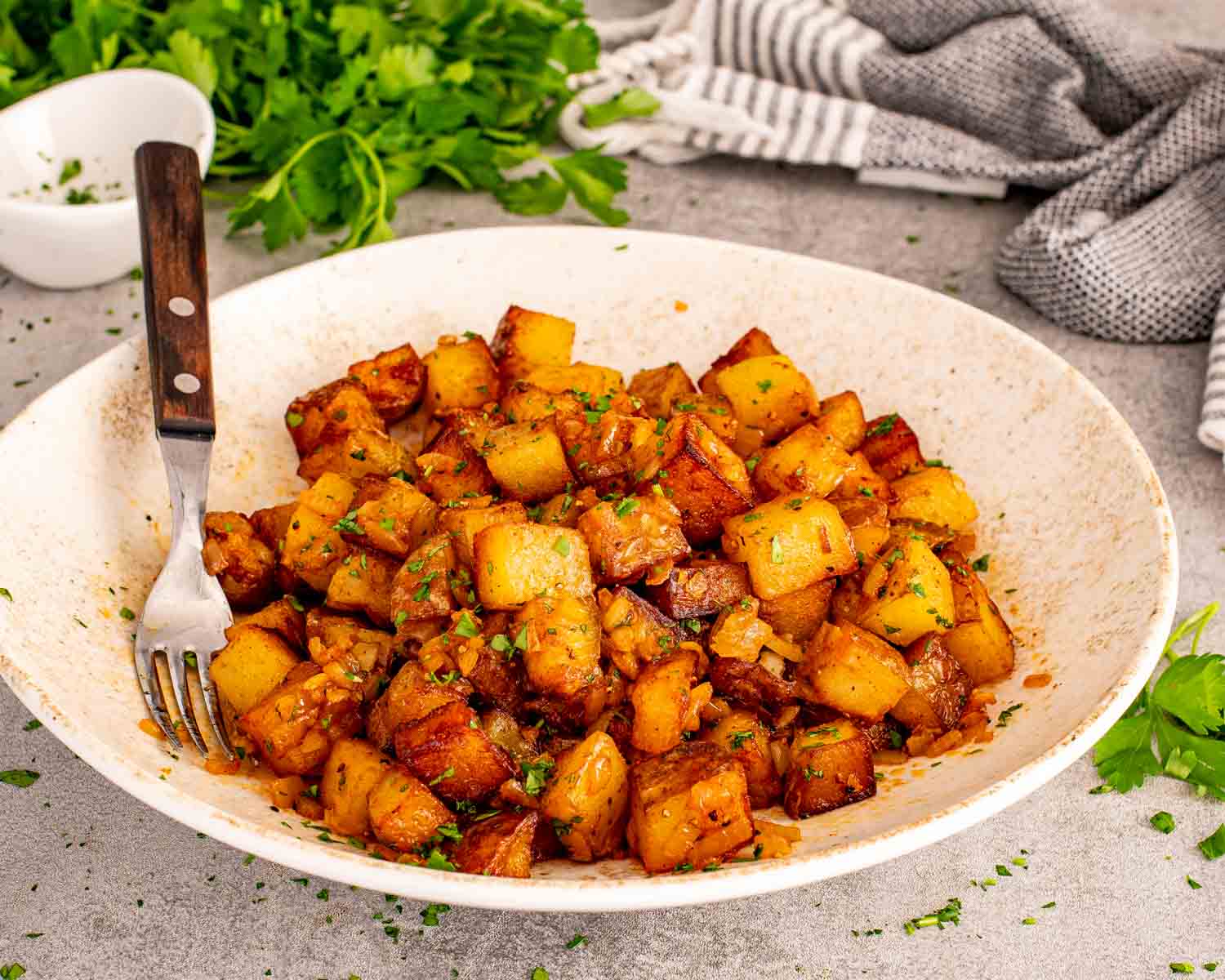 Close up rustic bowl filled with crispy home fries, caramelized onions, parsley, and golden browned potato cubes.