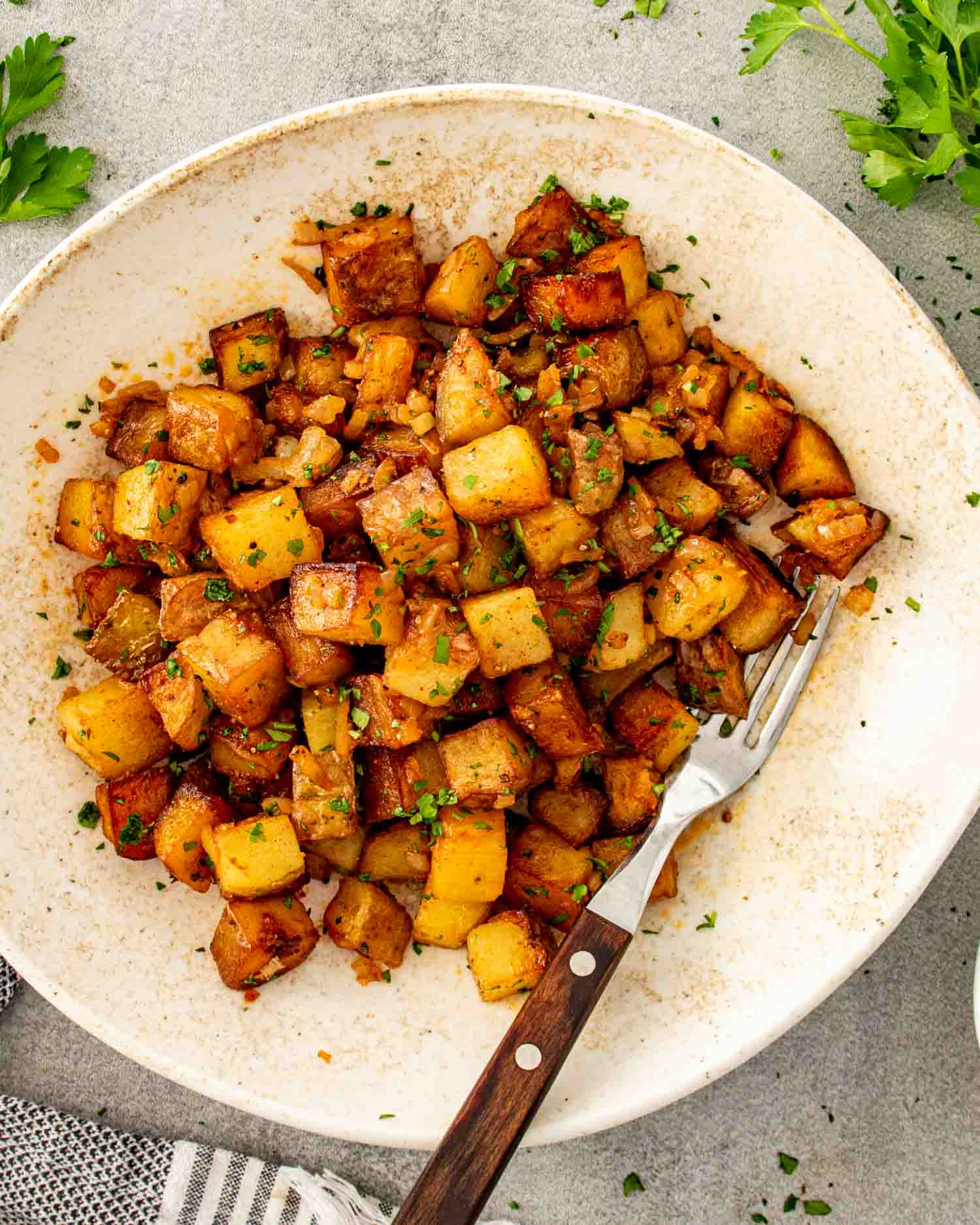 Crispy golden home fries served in rustic bowl with fork, parsley garnish, and cozy linen styling on gray surface.