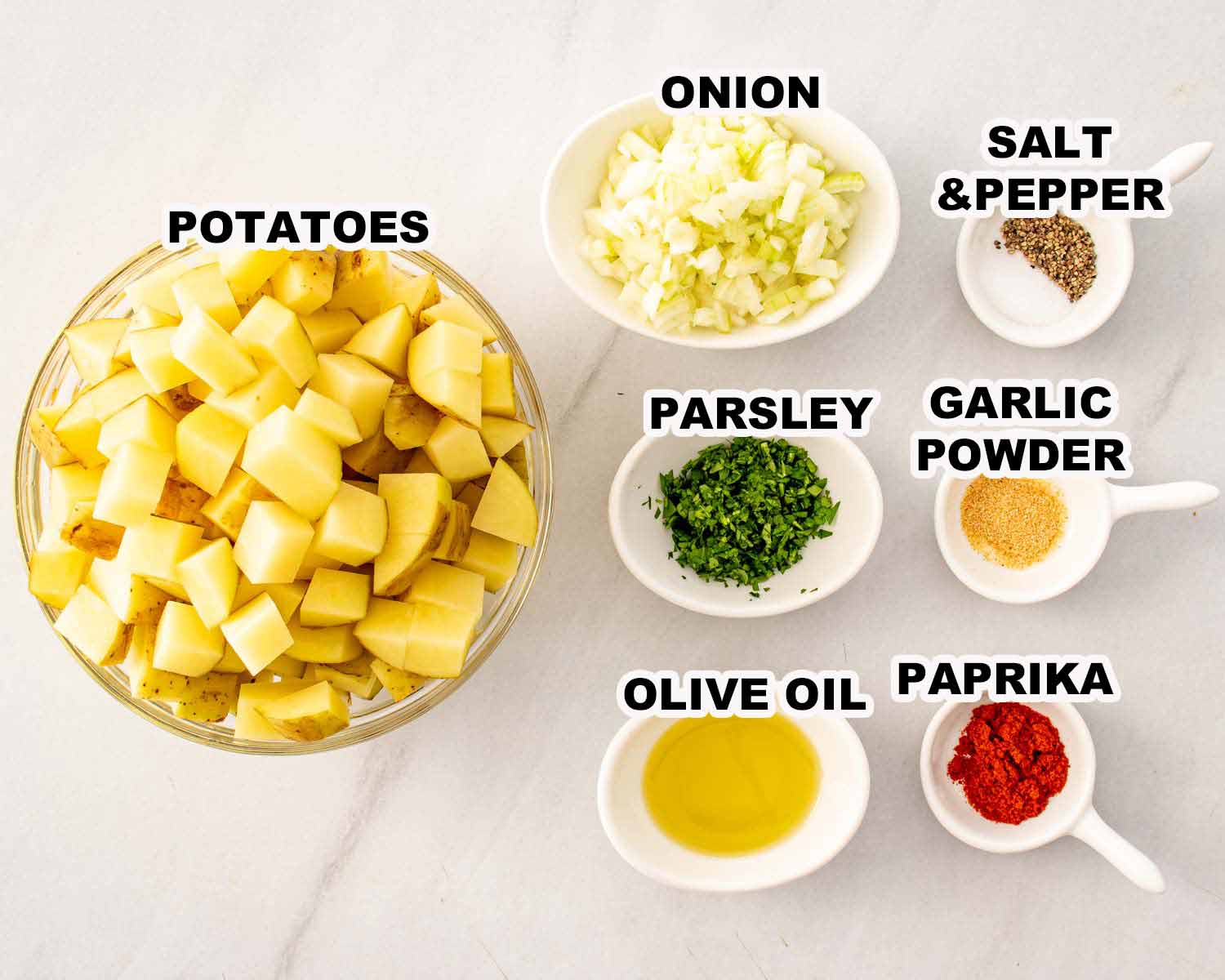 Home fries ingredients arranged in white bowls including potatoes, onion, parsley, paprika, garlic powder, olive oil, salt, and pepper.