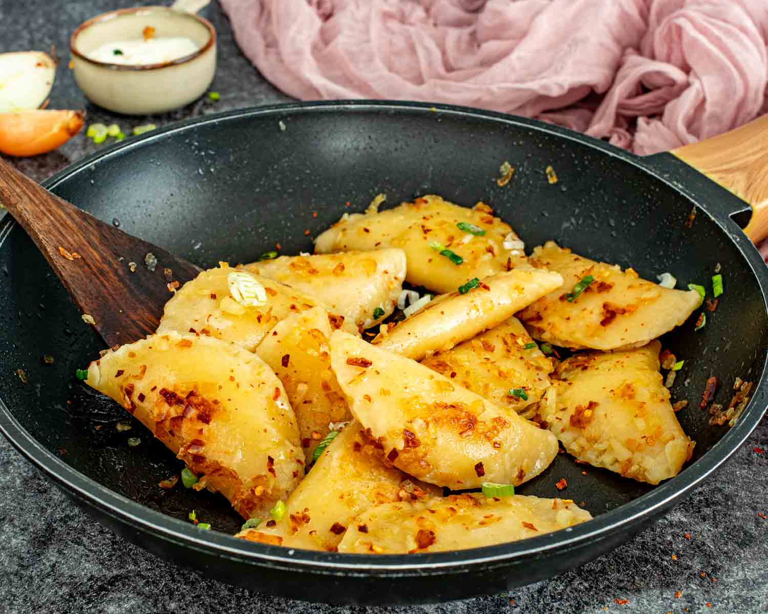 Golden pan fried homemade pierogi with caramelized onions and green onions being stirred with a wooden spoon in a black skillet.