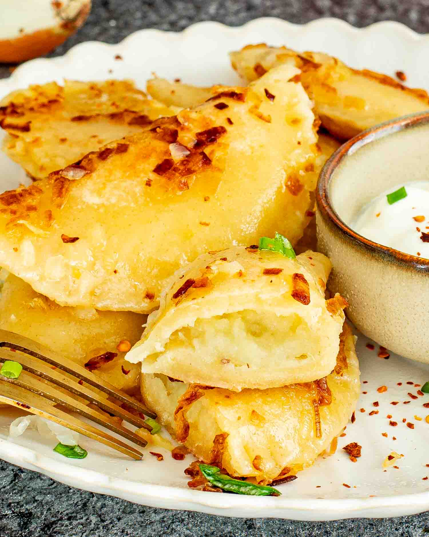 Homemade pierogi on a white scalloped plate cut open to show the potato cheddar filling with a small bowl of sour cream and a gold fork.