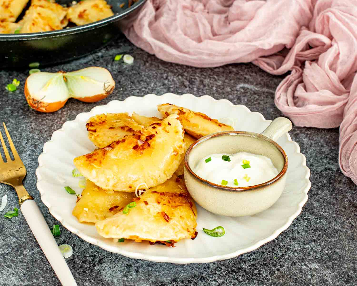 Golden homemade pierogi on a white scalloped plate served with a bowl of sour cream and chives with a pink linen and skillet in the background.