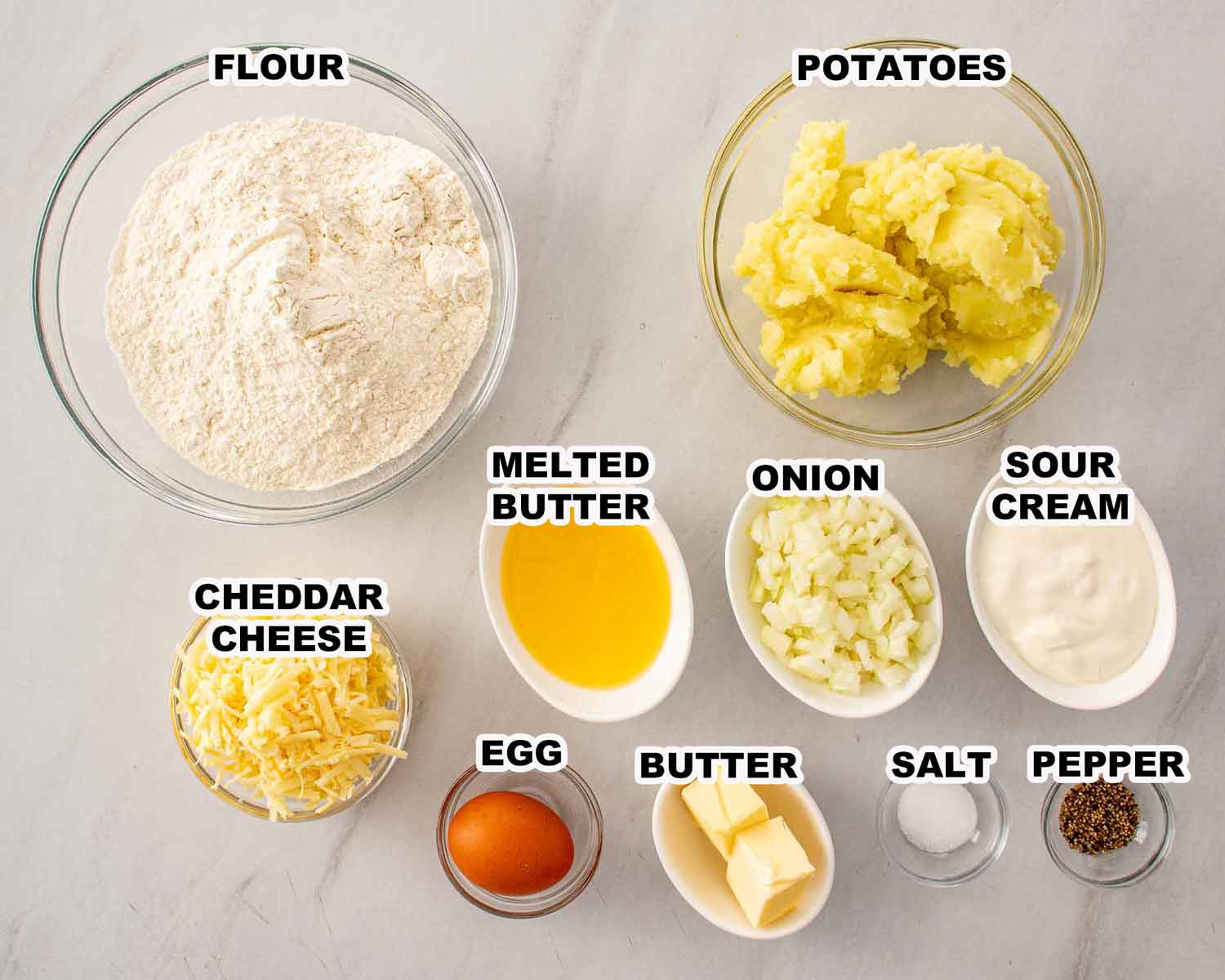 All purpose flour, mashed potatoes, cheddar cheese, egg, melted butter, onion, sour cream, salt and pepper ingredients for homemade pierogi laid out on a marble surface.
