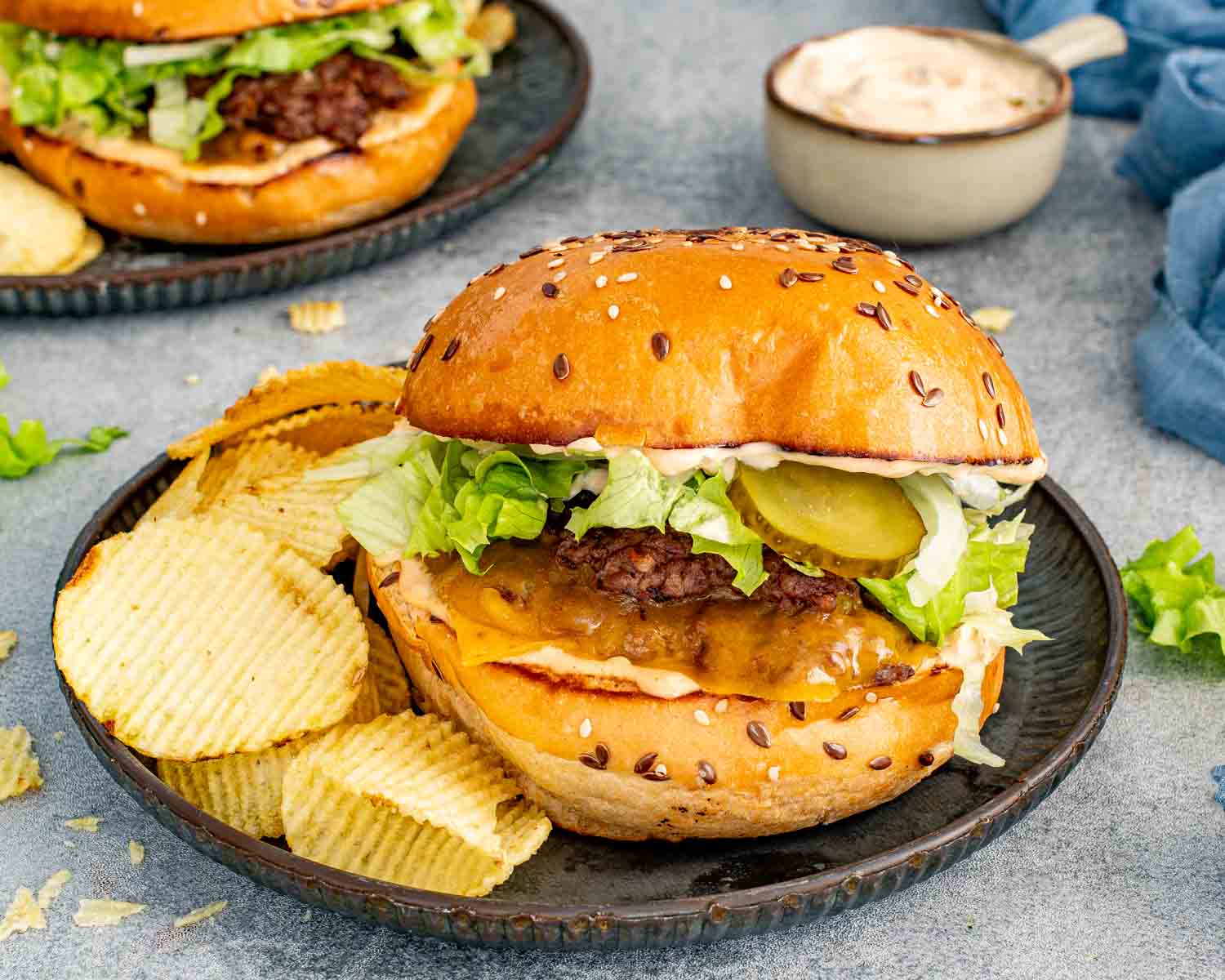 Two smashed burgers on a wood board with ridged chips, shredded lettuce, and a small bowl of special sauce in the background on a gray surface.
