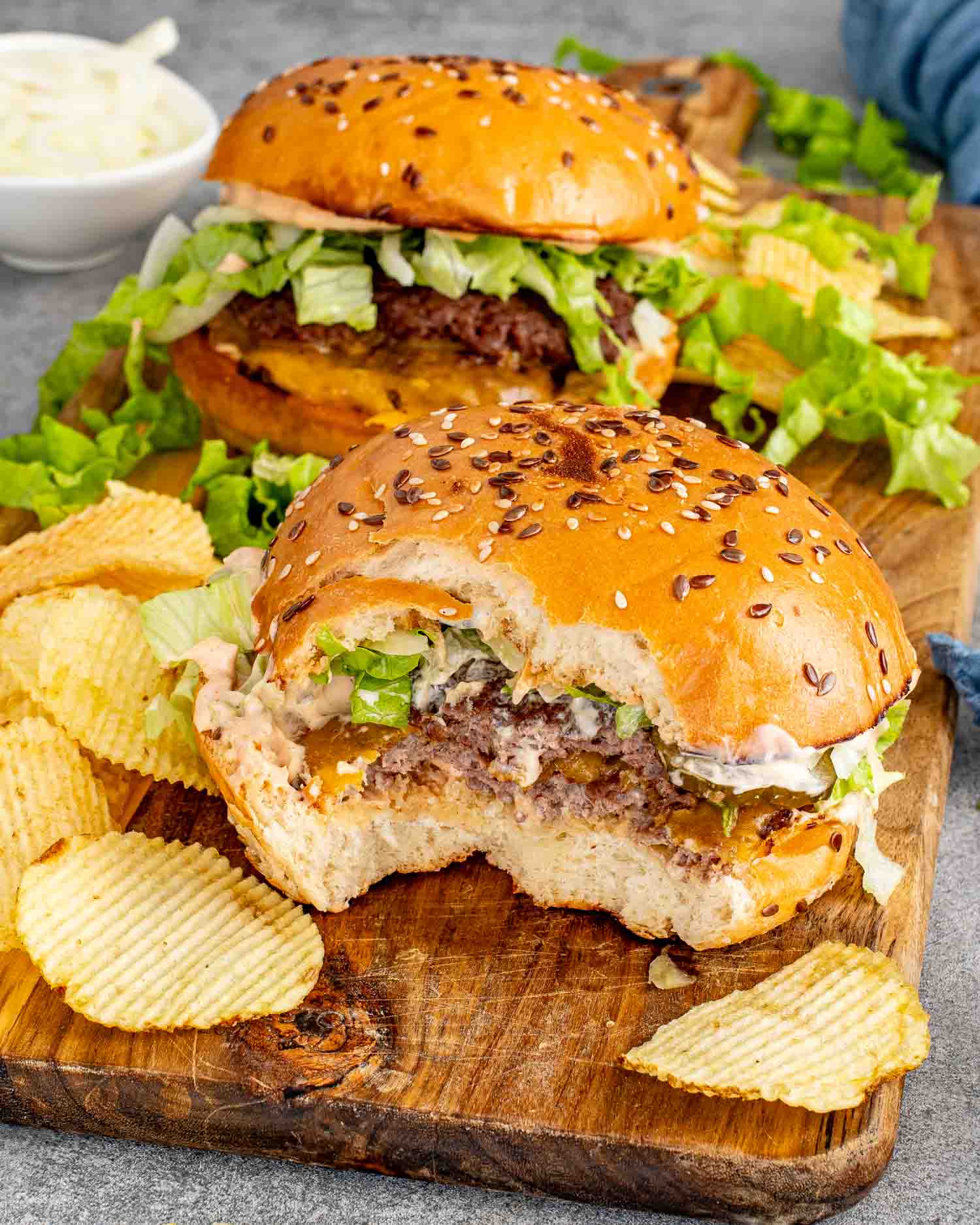 Close up of a smashed burger with a bite taken out showing the double patty, melted cheddar, special sauce, and shredded lettuce on a toasted sesame seed bun on a wood board.