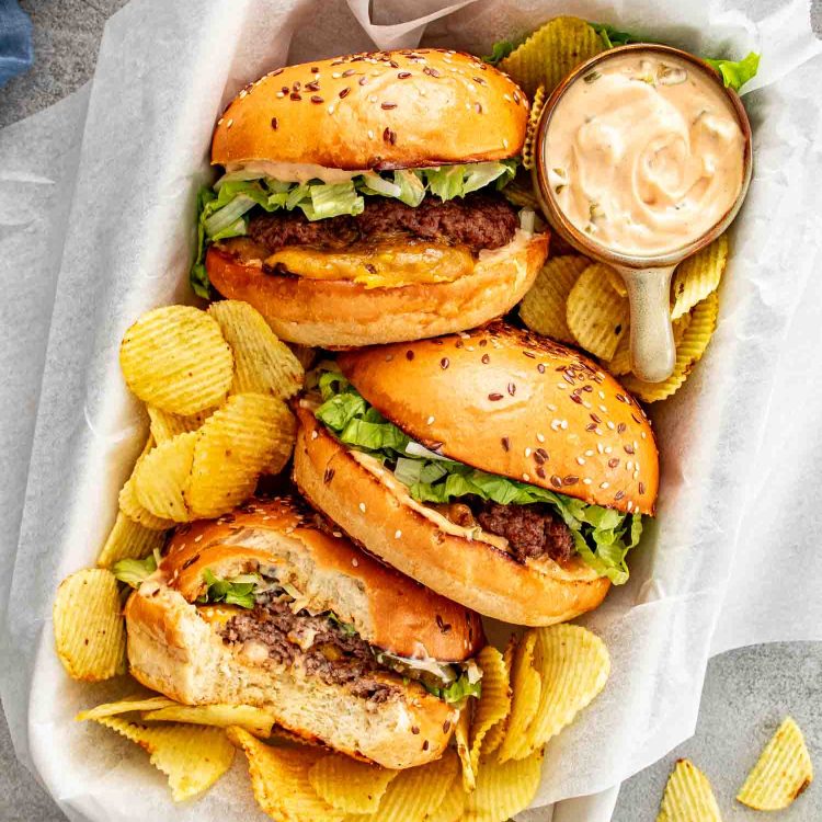 Overhead view of three smashed burgers in a parchment lined basket with ridged chips and a small bowl of homemade special sauce on the side.