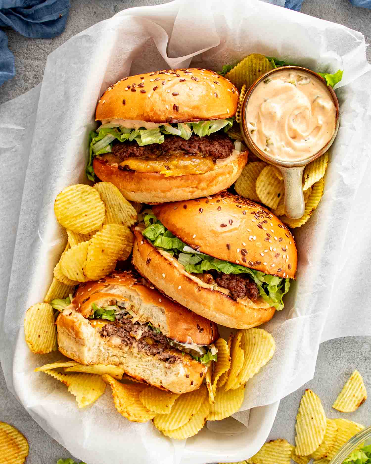 Overhead view of three smashed burgers in a parchment lined basket with ridged chips and a small bowl of homemade special sauce on the side.