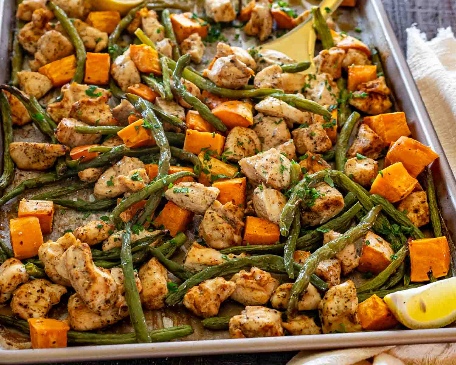 Close-up of roasted chicken bites, blistered green beans, and caramelized sweet potatoes on a parchment-lined sheet pan.