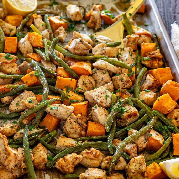 Golden roasted chicken, tender green beans, and caramelized sweet potatoes on a sheet pan with fresh parsley and lemon wedges.
