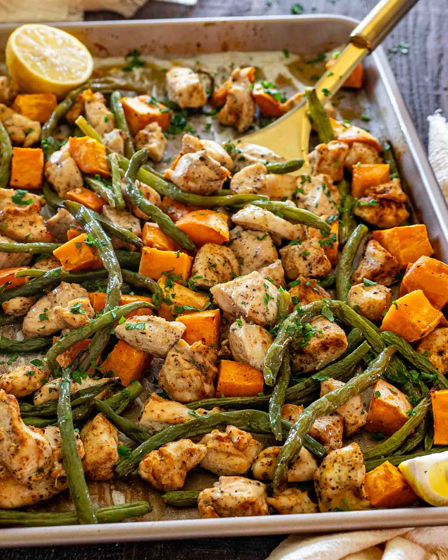 Golden roasted chicken, tender green beans, and caramelized sweet potatoes on a sheet pan with fresh parsley and lemon wedges.