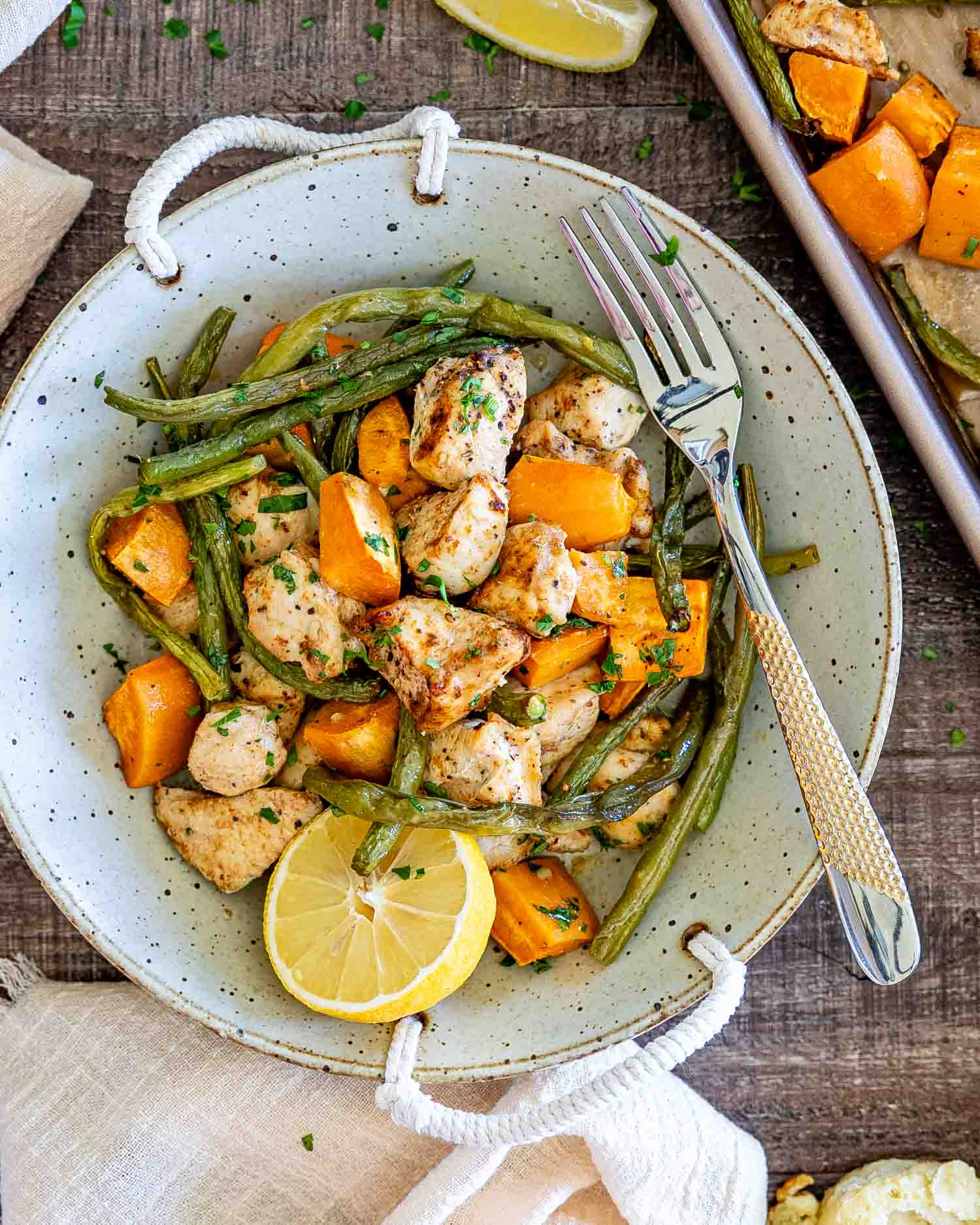 Bowl filled with roasted chicken, green beans, and sweet potatoes, garnished with parsley and fresh lemon beside the sheet pan.