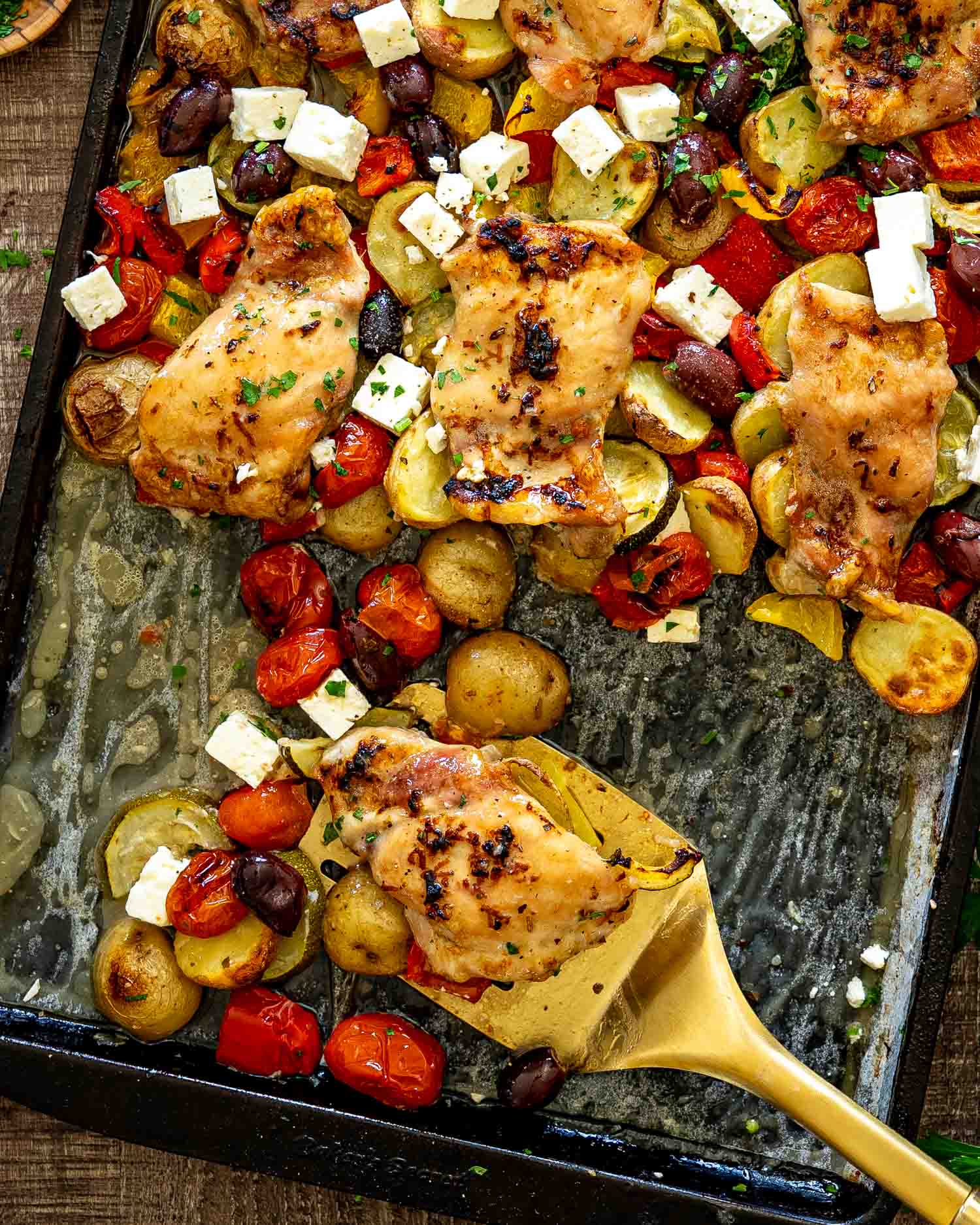 Serving spatula scooping Greek chicken and vegetables from sheet pan loaded with feta, olives, potatoes, and tomatoes.