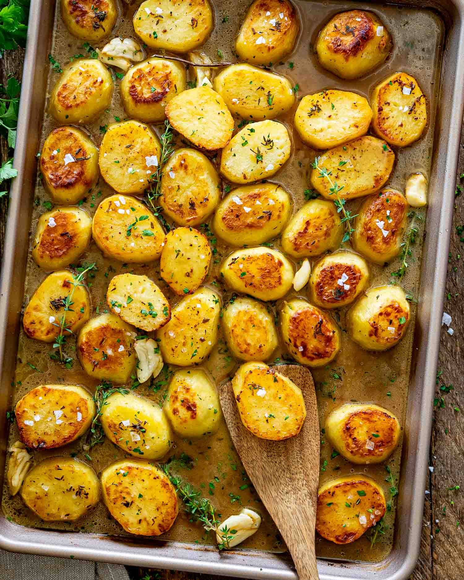 Overhead view of crispy melting potatoes in buttery broth with garlic, thyme, and wooden spatula lifting one perfectly browned potato.