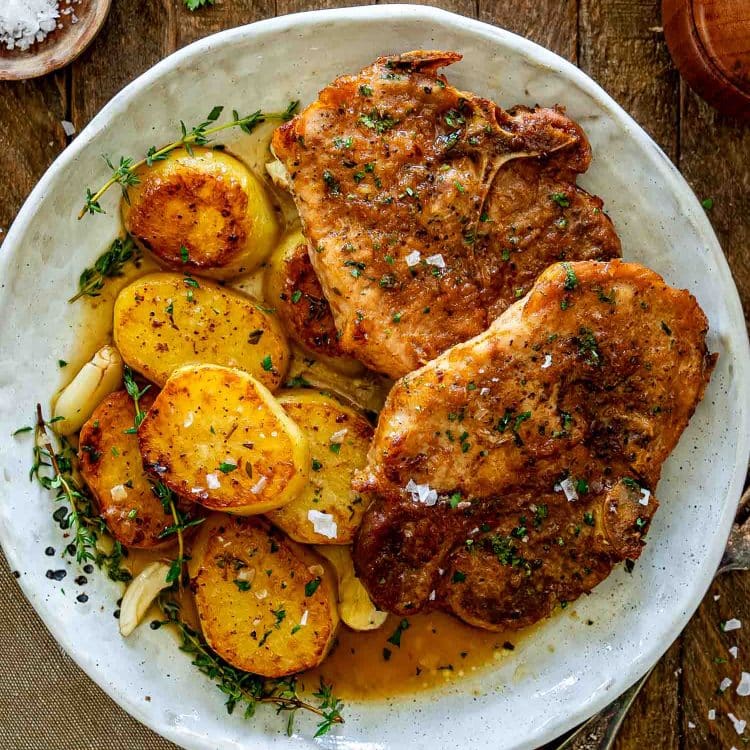 Honey garlic pork chops served with roasted potatoes, fresh thyme, garlic cloves, and savory pan juices.