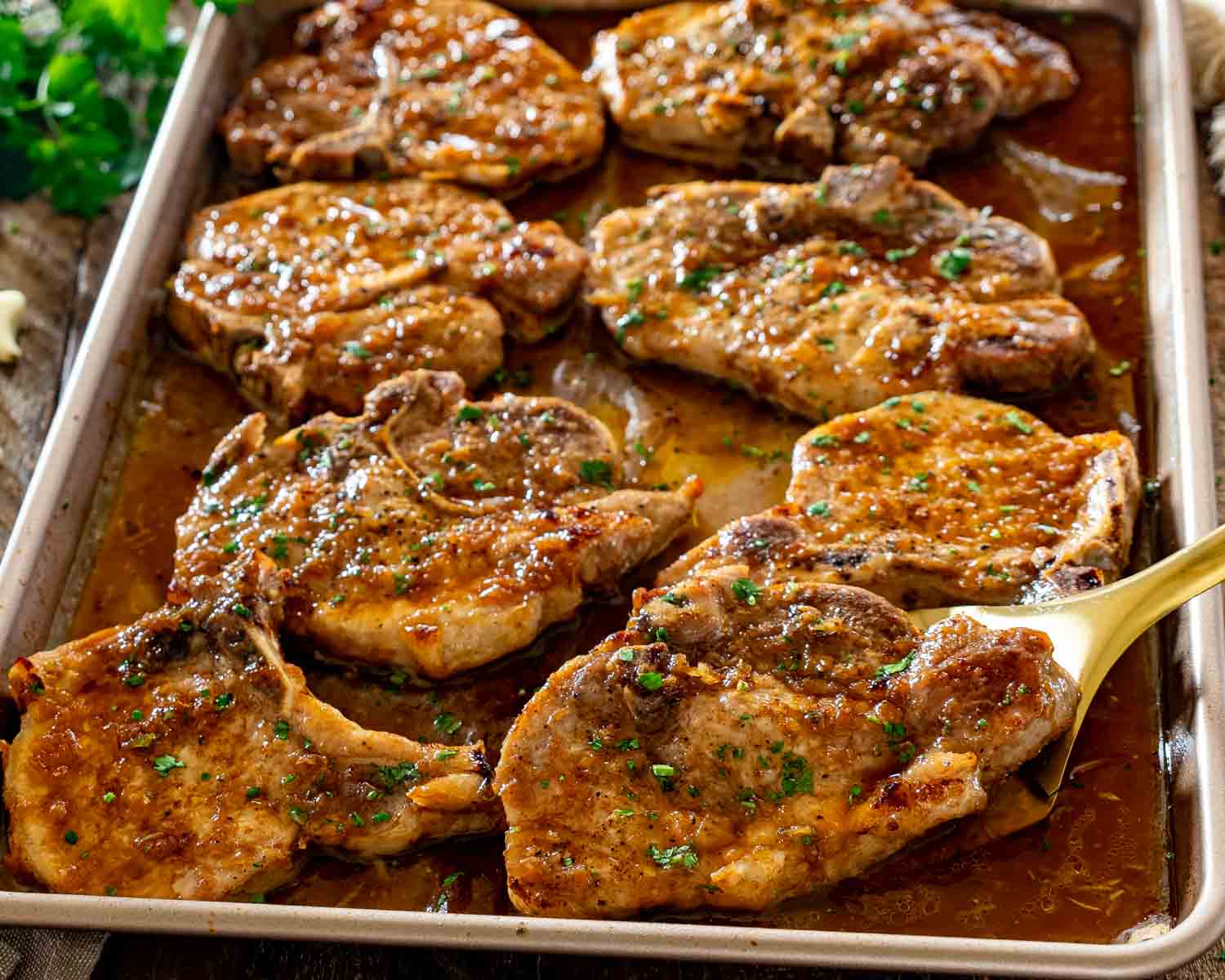 Glazed honey garlic pork chops resting in rich pan sauce on baking sheet with serving spatula underneath.