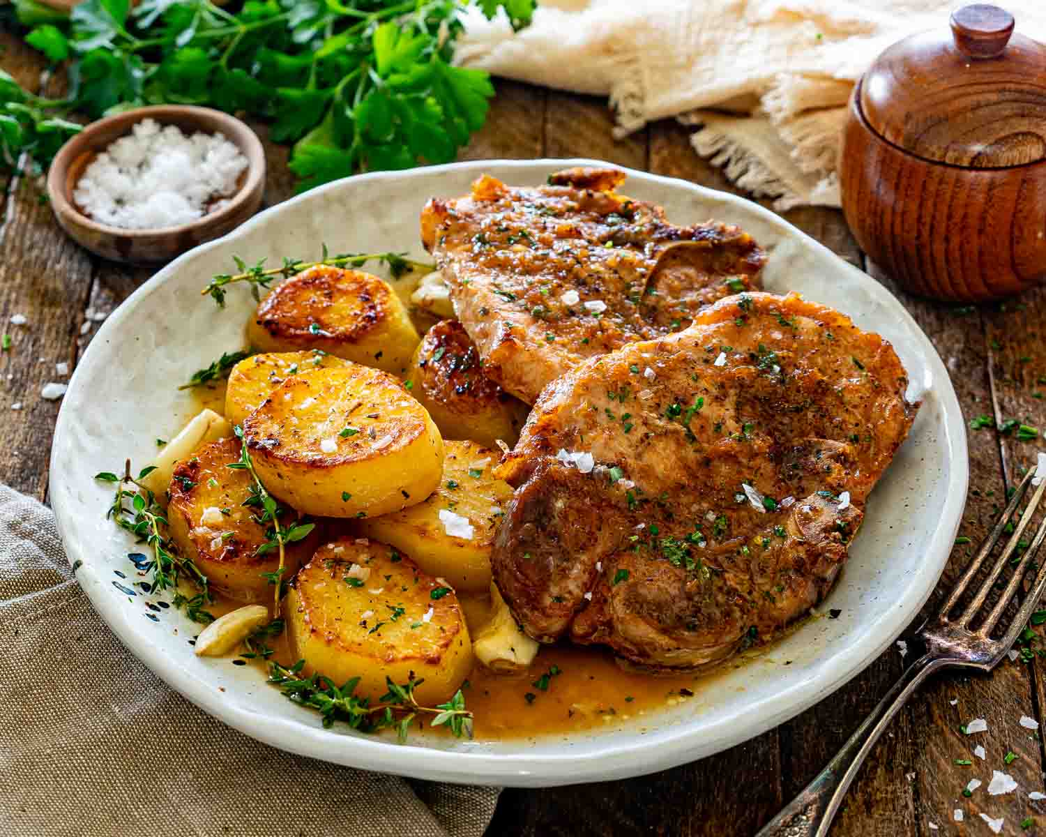 Two juicy honey garlic pork chops with crispy roasted potatoes and herbs served on rustic ceramic plate.