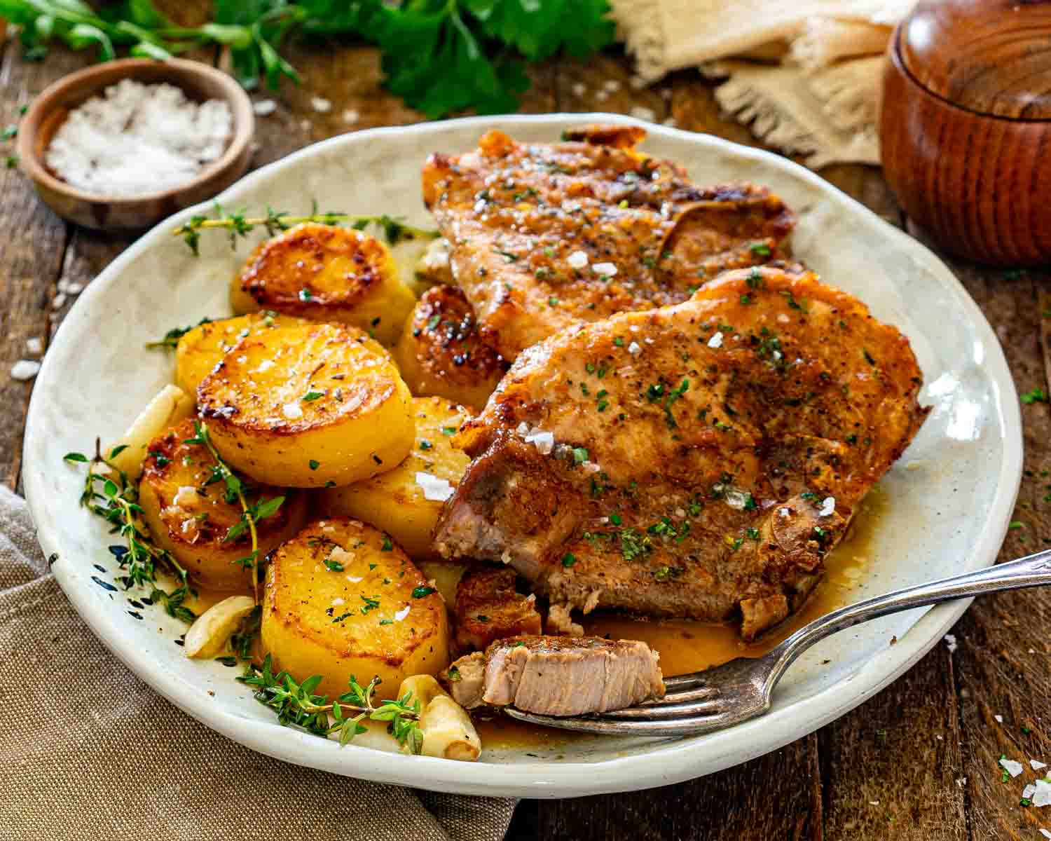 Two juicy honey garlic pork chops with crispy roasted potatoes and herbs served on rustic ceramic plate.