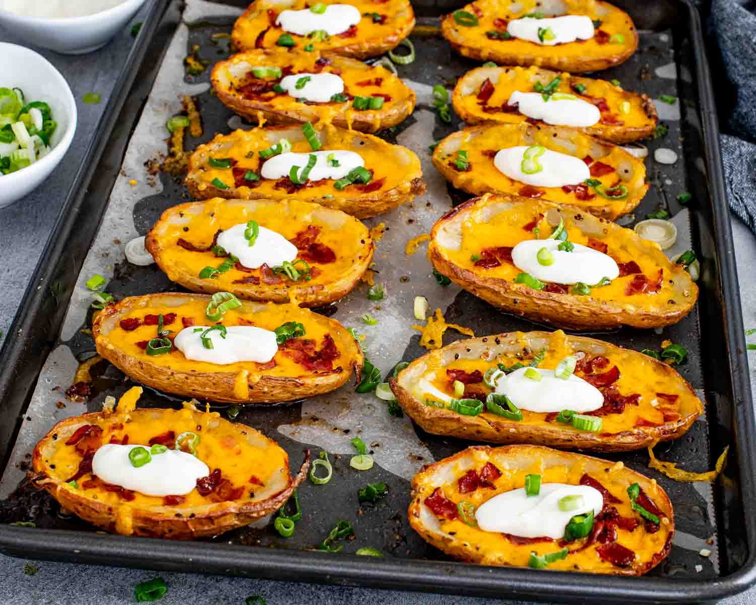 12 crispy potato skins loaded with melted cheddar cheese, crumbled bacon, and sliced green onions fresh out of the oven on a dark baking sheet.