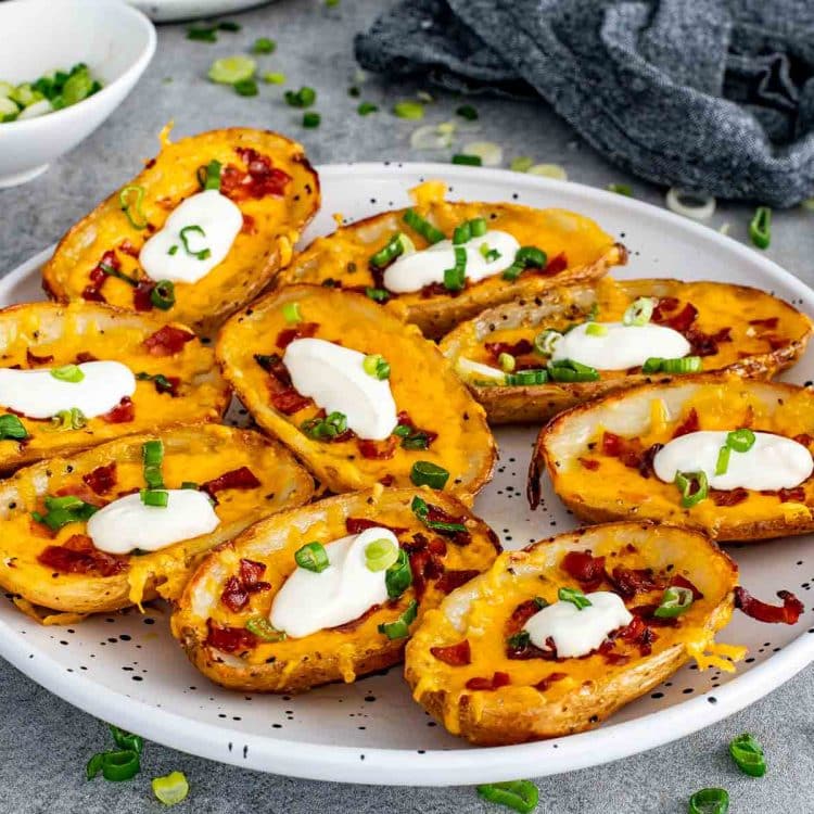 A full plate of crispy restaurant style potato skins loaded with melted cheddar cheese, crumbled bacon, green onions, and dollops of sour cream.