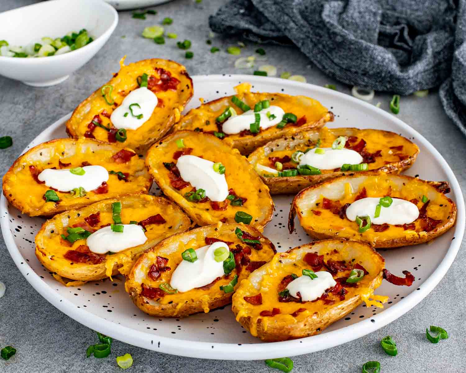 A full plate of crispy restaurant style potato skins loaded with melted cheddar cheese, crumbled bacon, green onions, and dollops of sour cream.