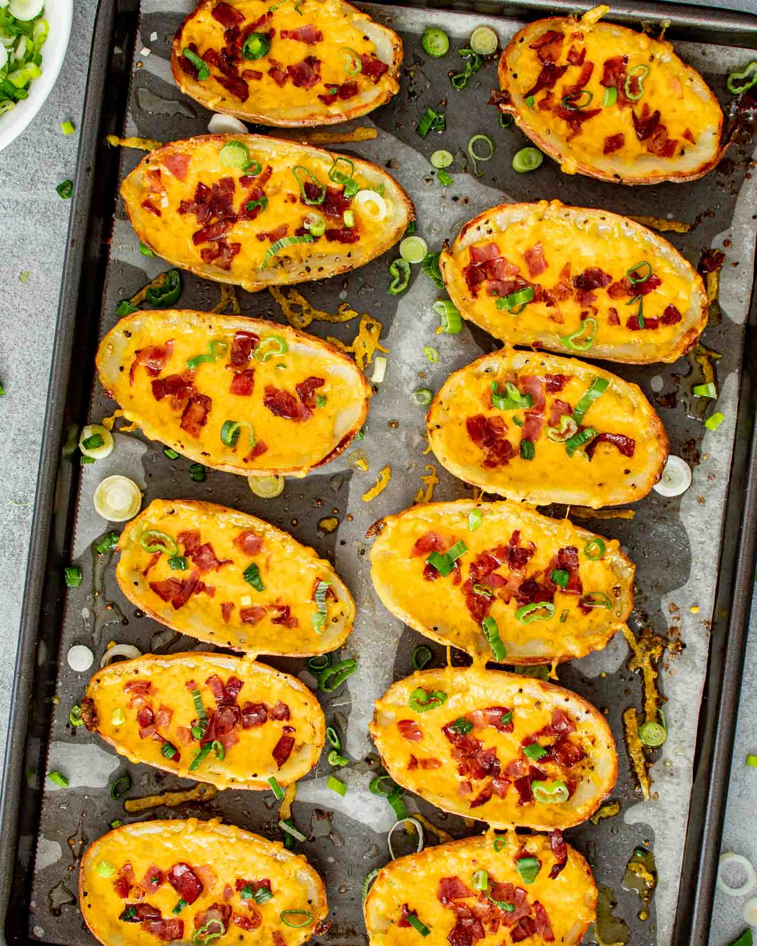12 crispy restaurant style potato skins topped with melted cheddar, crumbled bacon, green onions, and dollops of sour cream on a dark baking sheet.