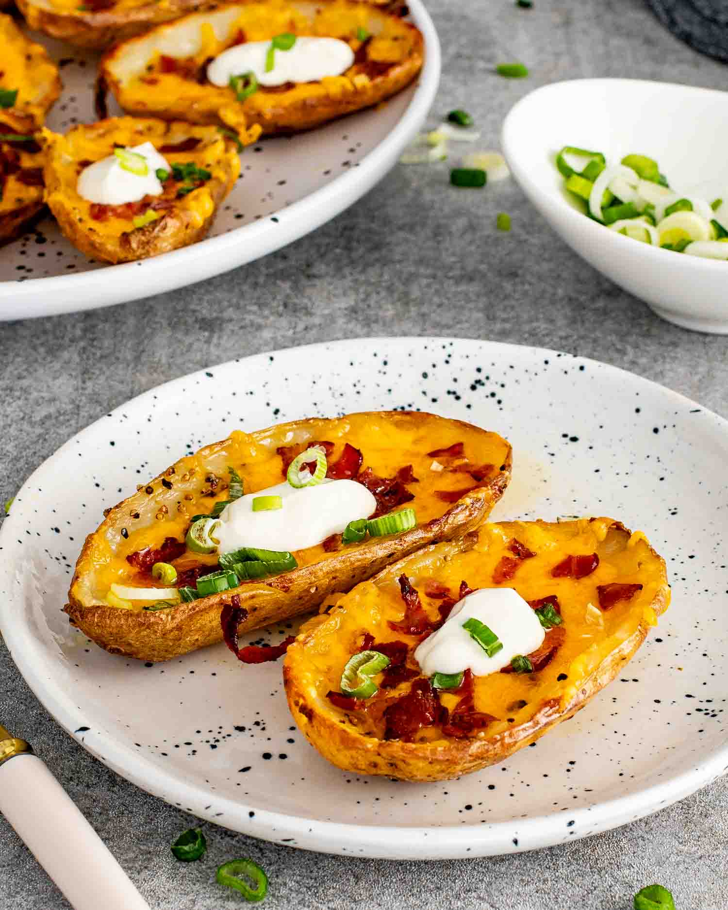 2 crispy restaurant style potato skins loaded with melted cheddar, crumbled bacon, green onions, and a dollop of sour cream on a white speckled plate.
