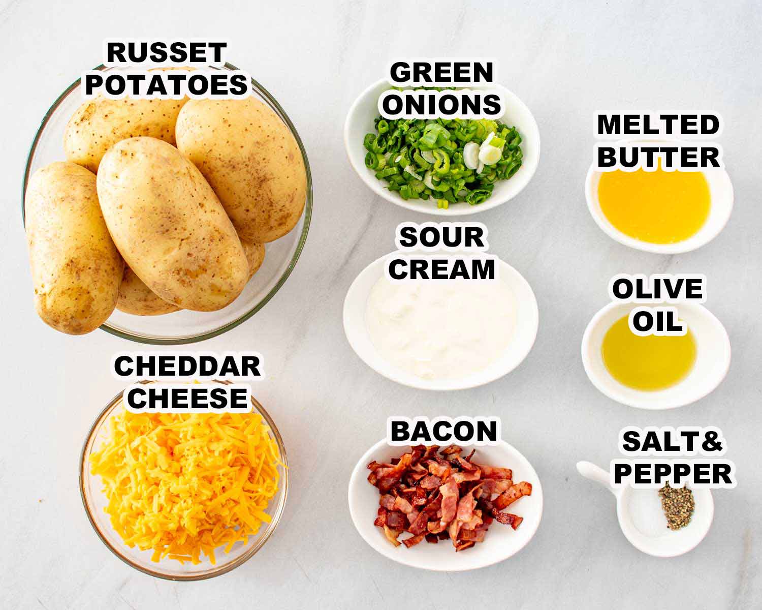 All ingredients for crispy potato skins laid out on a white surface including russet potatoes, cheddar cheese, bacon, green onions, sour cream, melted butter, olive oil, salt and pepper.