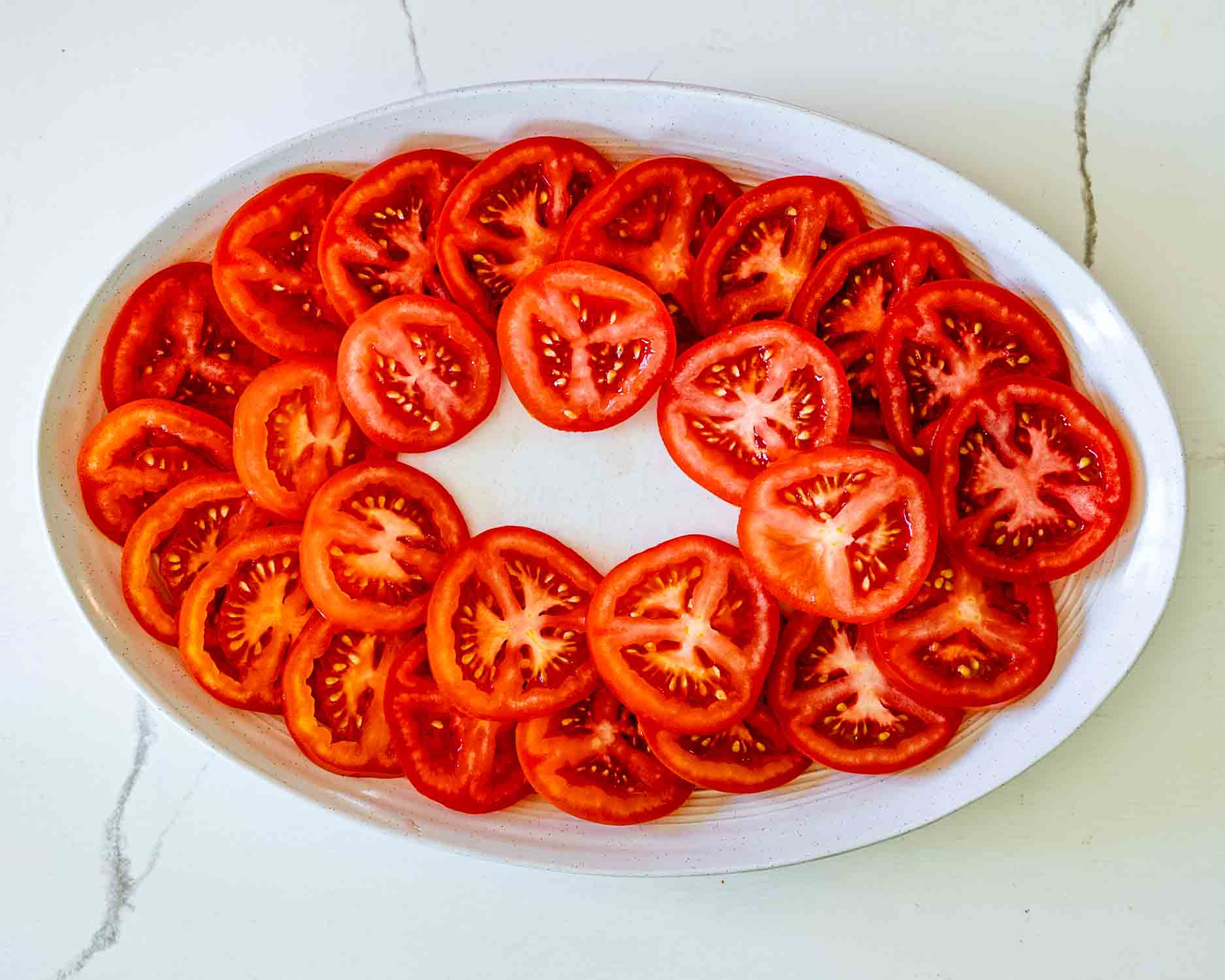 Overhead view of fresh sliced tomatoes neatly arranged on white serving platter for tomato burrata salad preparation.
